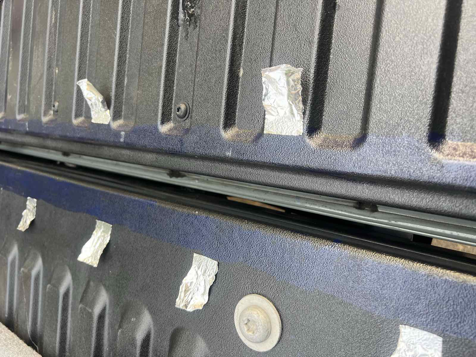 Squares of aluminum foil tape stuck to a Tacoma bed floor and tailgate to mask the ends of an adhesion promoter line before the ROK Block seal is applied.
