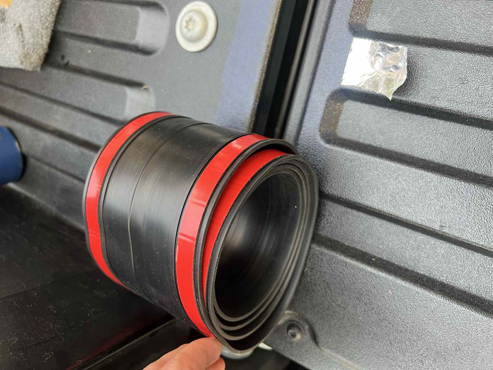 The very start of a ROK Block tailgate seal being applied at the left end of a Tacoma bed floor, with the red adhesive backing partially peeled.